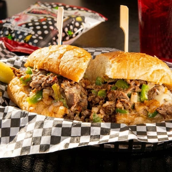 Cheesesteak sandwich with green peppers, onions, and melted cheese, served with a pickle and drink.